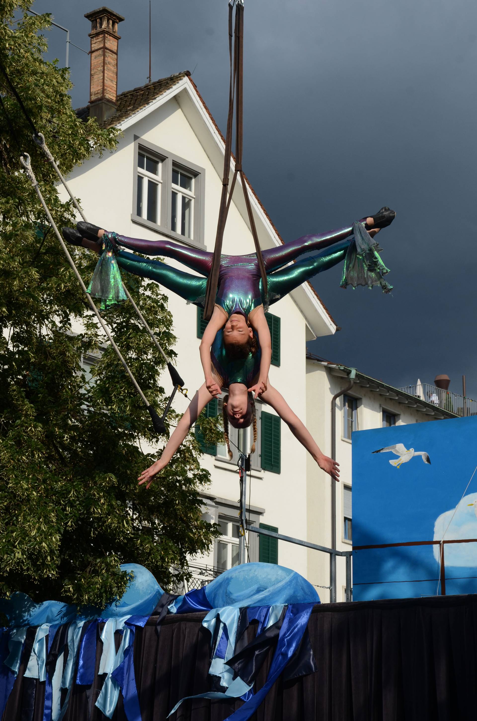 An acrobatic performance by the children's team of Kinderzirkus Robinson in Switzerland.  Two girls are hanging up side down from a bungee doing the spllits in the air.  They are both wearing shiny costumes.  