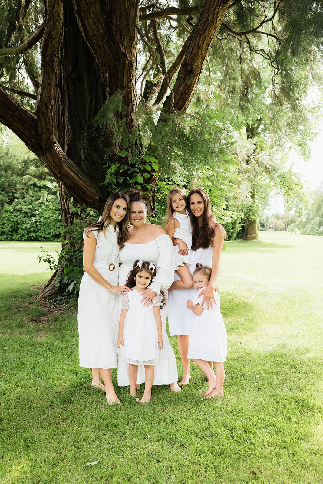 A family photo of all the generations of women in a family in the coutryside in a lush green lawn with a large tree in the background.  All ladies and girls are wearing white and are captured by Mary Fernandez, family photographer in Switzerland. 