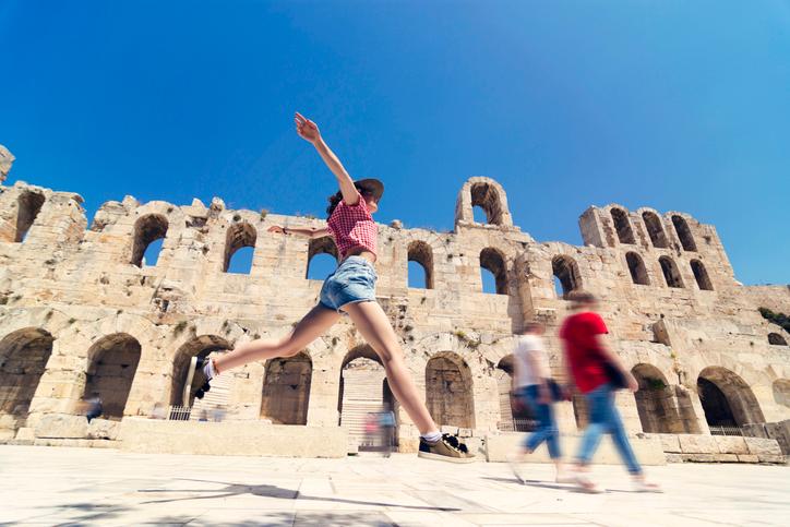 A young teenage girl in shorts, t-shirt and a summer cap taking a big leap in front of the ancient Roman Theater Herodion in the heart of Athens, Greece.  One of the many interesting sites to visit while traveling to Athens with kids.  