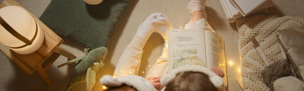 A young toddler girl in white socks and pyjamas is on a white carpet the floor with her sibling reading a book from the book club.  They are surrounded by fairy lights and they are reading a story about acts of kindness and giving.  