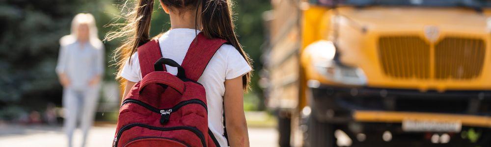 A primary school girl in pig tails and a read backpack is heading to school, and is having a difficult time separating from her mother, but must go ahead anyway.  