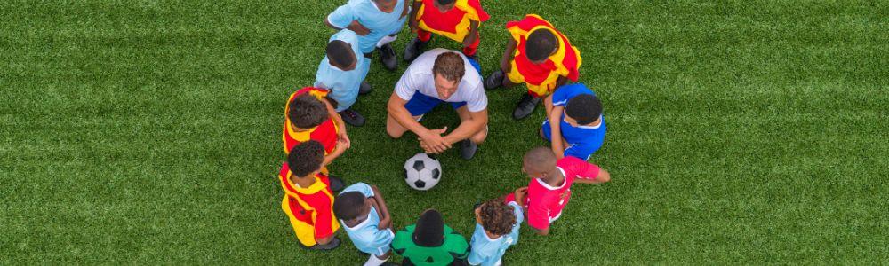 A young male soccer couch surrounded by 12 pre-teen boys in a circle while he is coaching them how to play in teams on a grass field in Switzerland.  