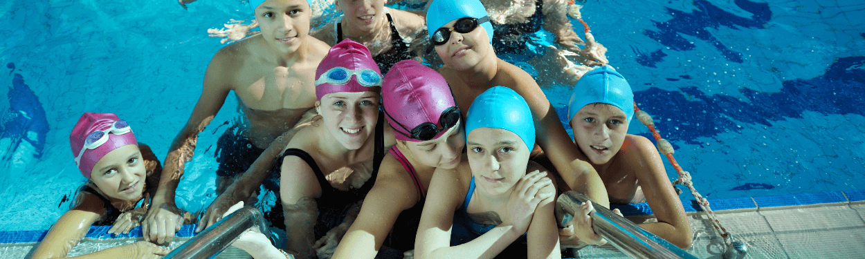 A swimming class of kids and pre-teens poses for a group photo inside the pool, with their swimming caps on