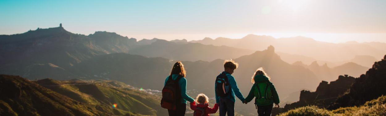 A group of children  of all ages, including a toddler, have arrived to the peak of the mountain in the Swiss Alps and are enjoying the view and the sunset holding hands. They are prepared for an overnight stay at a mountain hut. 