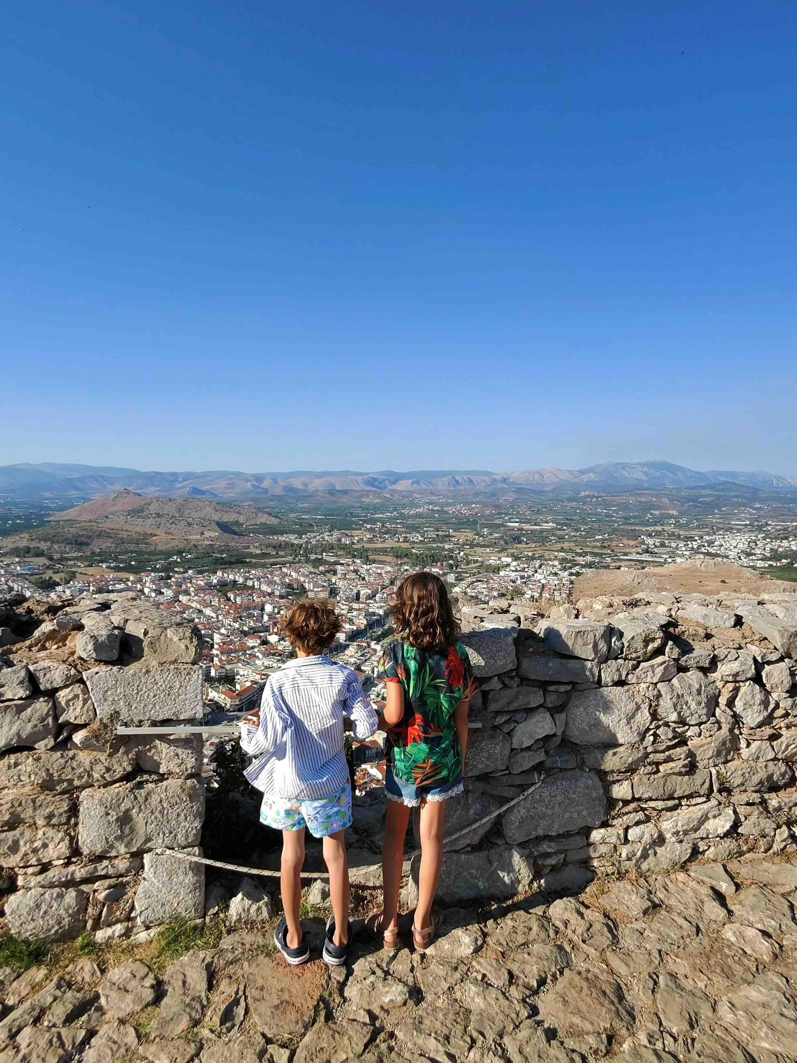 A small boy and girl wearing summer clothes are observing the view of the city of Nafplio from the top of Palamidi Castle, under the summer sun in Greece.