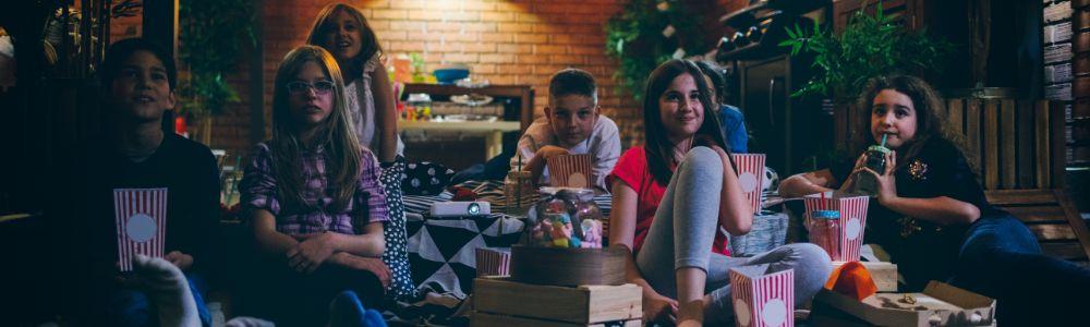 A group of pre-teens gathered together in a home-garden enjoying popcorn while watching a screening of a favorite movie outdoors.  It looks like a celebration with pizza and soft drinks on a summer's evening.  