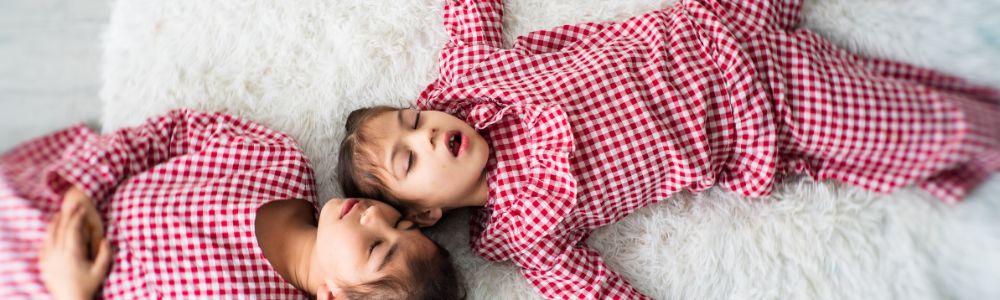 Two schoolgirls in matching checked white and red long pyjamas sleeping with their mouths open on a white fluffy carpet.  