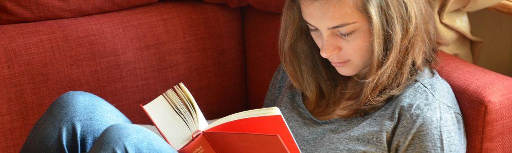 A teenage girl is snuggled on a red sofa reading a red hard-covered book.  She is enjoying it and is absorbed by the story.  