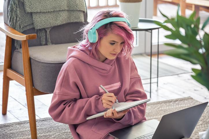 A trendy teenage girl with headphones taking notes and doing her homework online