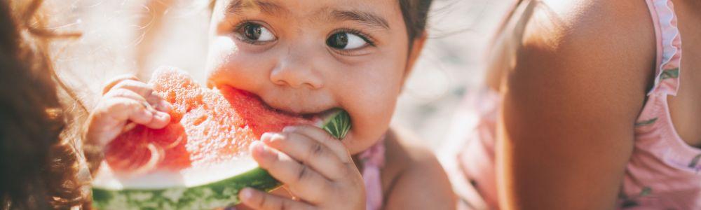 Close-up of a toddler girl in a swimsuit muching on a slice of watermelon at the restaurant Cheimonas Kalokairi sti Zouberi, in Nea Makri, in Greece.