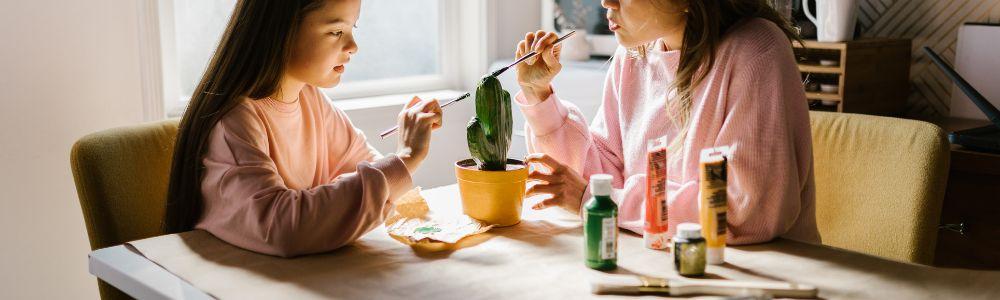 A mother and school-aged daughter are sitting together and chatting while paiting a cactus sculpture in a white sun-filled room.  Art is a perfect way to connect with young children and find opportunities to begin talking and bonding.  