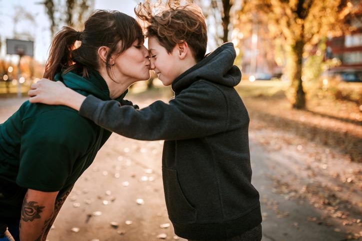 A mother and teenage son in the middle of the street.  They are embraced and exchanging a loving kiss by the mother on her son's nose.  He is very happy.  