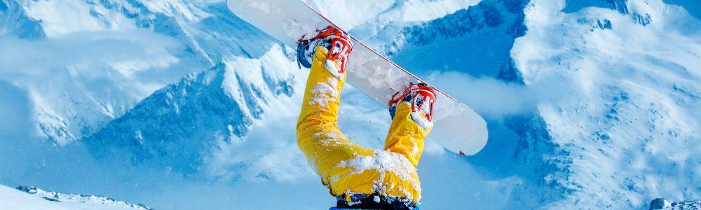 A photo of a snowboarder in yellow ski-pants, with is feet and snowboard up in the air, posing funnily and doing childs play with his head in the snow.