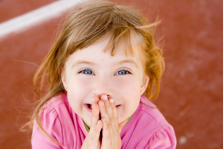 A small red-haired girl aged 6 or 7 years old is smiling covering her mounth, as if to show she is telling a little lie and can't help laughing.