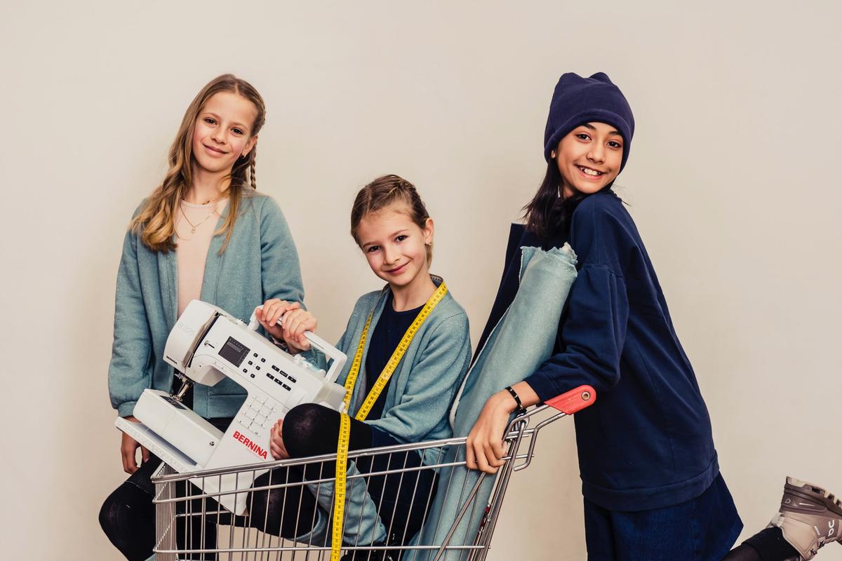 Three teenagers in light jean shirts, posing together, while one is in a supermarket cart with a sewing machine, fabric and a measuring tape, all ready to start sewing and designing clothes at Allegra Fashion Design Camp in Zollikon, Switzerland.  