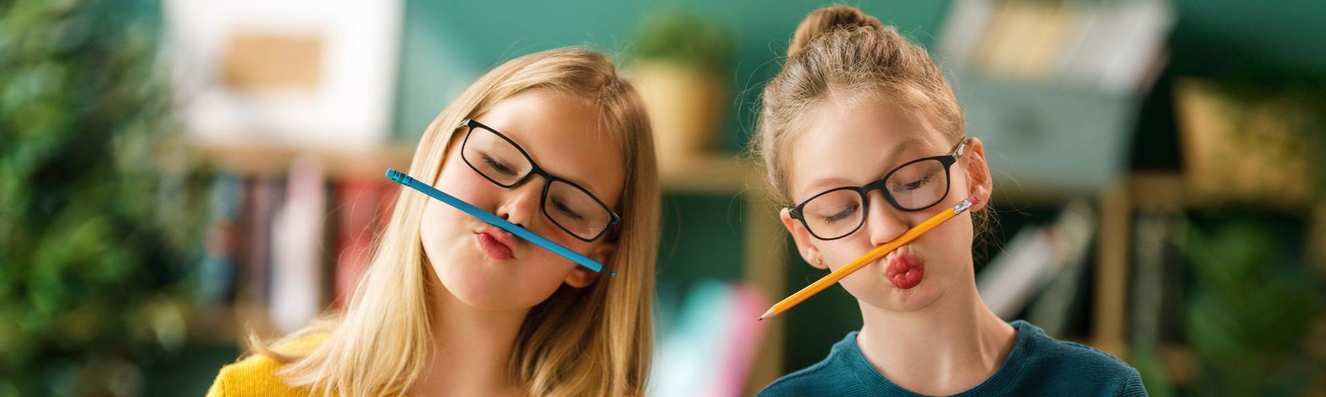 Two pre-teen girls are posing in their English class at AtoZ English with pencils between their nose and lips.  They are wearing eye-glasses and closing their eyes to keep their pencil tightly held between their nose and and lips.  