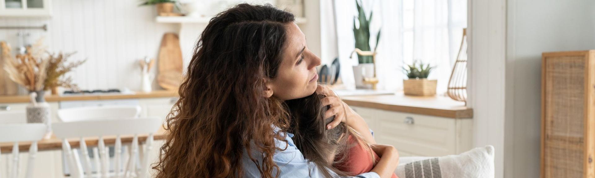 A white brown-haired mother embracing her pre-teen daughter on the sofa of their sunlit house, comforting her before her first day of school which is bringing her anxiety.  They are by the kitchen and can potentially have a comfort snack together.  