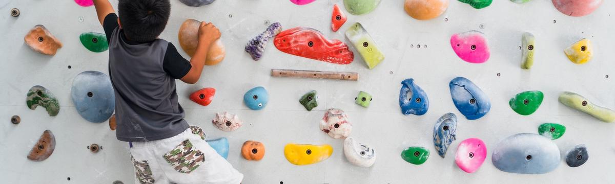 Young child climbing an indoor bouldering wall with colorful handholds and footholds, viewed from behind.