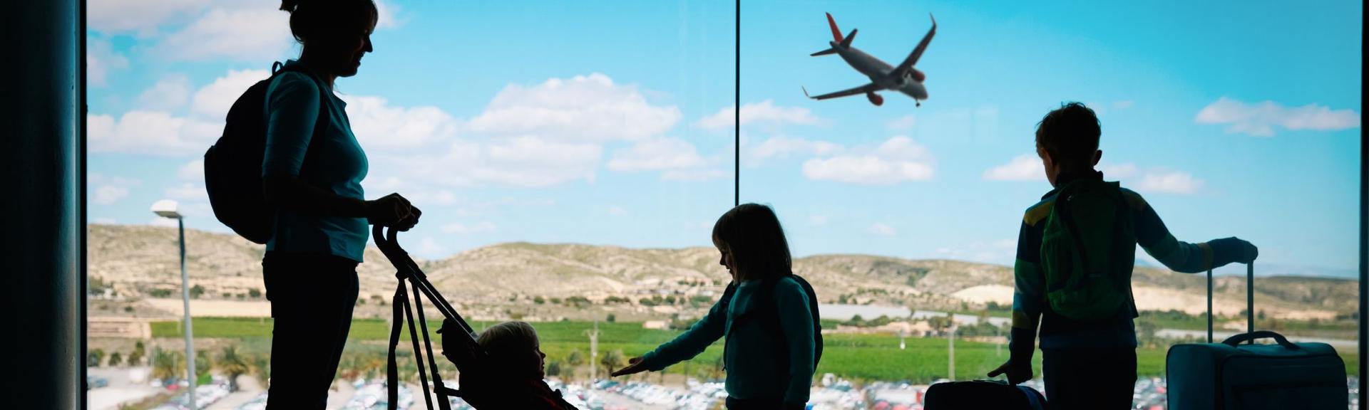 A mother and her three children by a floor to ceiling airport window getting ready to travel.  They are prepared with board games in their backpacks and games to keep them busy during their trip.  One child is in a buggy and the other two are standing nearby looking out the window holding by their luggage.