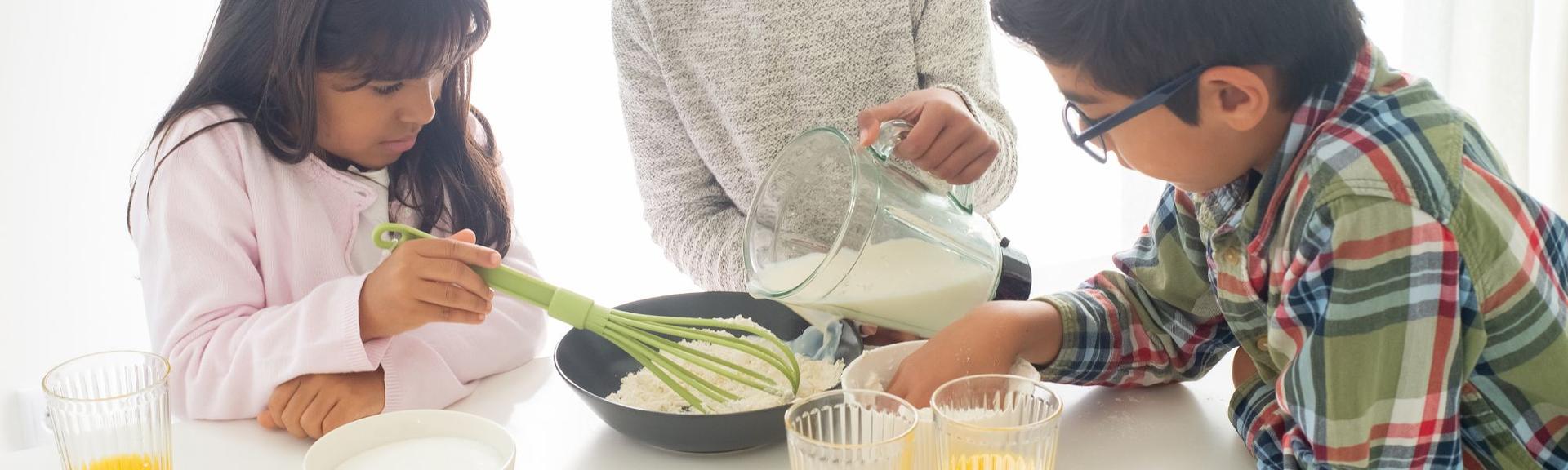 A group of 3 pre-teens are working together to prepare a mix for baking on a white table and white background.  One girl is pouring milk from a measuring pitcher in a bowl of flour while the second girl is ready to mix and the boy is measuring the next ingredent.  