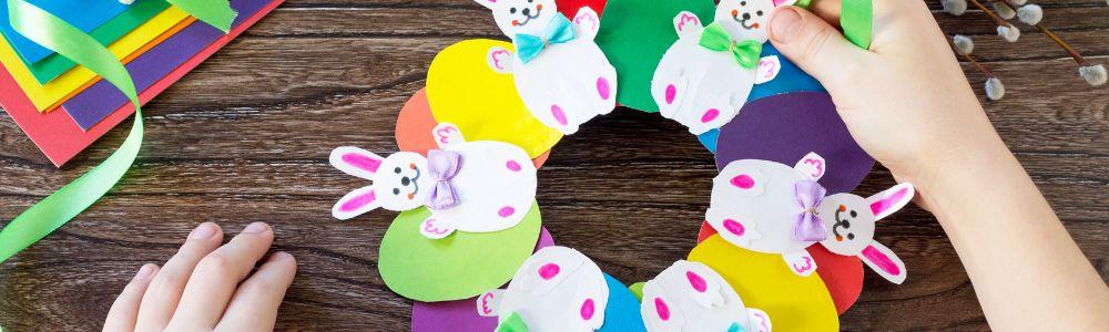 A colorful Easter wreathe on a wooden table. It is made out of cut-out paper in shapes of colorful Easter eggs and white Easter bunnies with a colored bow tie.  Paper and crafts for Easter decor is spread around the wooden table.  