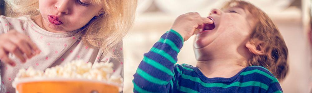 Two young siblings are eating popcorn together while getting ready for a family movie night at home.  