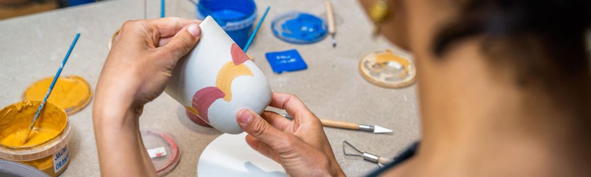 In a children's pottery class at the Gas Museum in Athens a child is holding a ceramic cup and painting in with pink and white curves and brushes.  