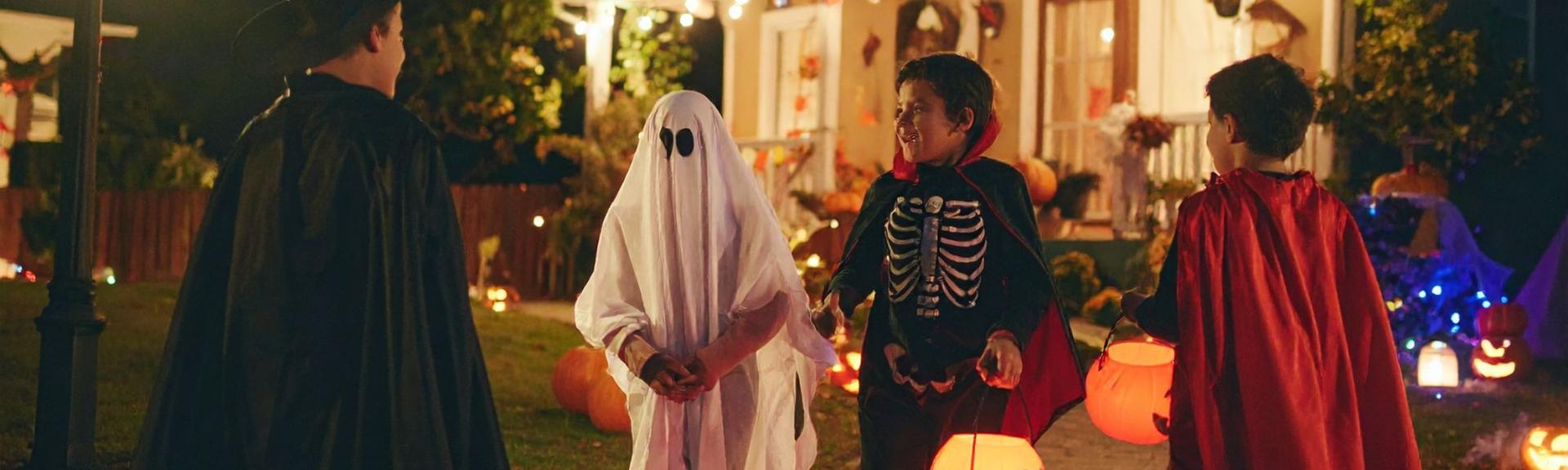 Four teenage boys in halloween costumes are walking around their neighborhood trick or treating, there is one with a ghost costume, one with a skeleton, one with a red cape and the fourth boy is wearing a black cape.  They are holding halloween lanterns with candles inside 