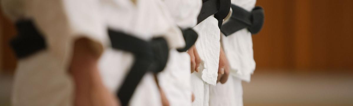 A close-up of several individuals standing in line wearing traditional martial arts uniforms (likely karate gi) with black belts, indicating advanced rank. The photo is focused on the waist area and the black belts, suggesting a formal or disciplined setting, possibly before or after a practice or grading.