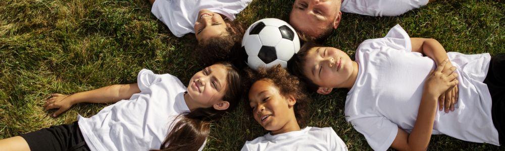 A group of young teenage boys and girls in white t-shirts and black shorts are lying on a grass soccer field with all their heads in a circle around a football, smiling happily tired, at an Intersoccer Birthday party near Zurich and Zug, Switzerland.  