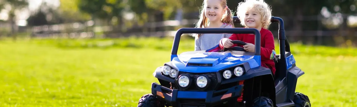 Two smiling children riding in a blue electric toy car on a sunny grassy field, enjoying an outdoor driving activity.