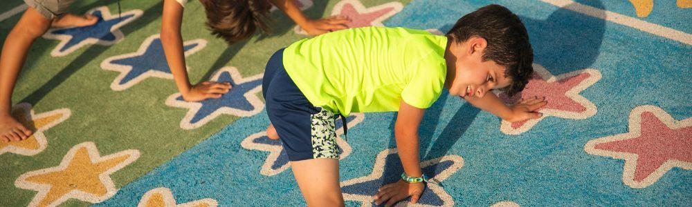 A school boy with a yellow fluorescent t-shirt is on a twister mat with stars playing touch the color. He has his hands on the floor and trying to balance along with another school girl his age. They are playing together and learning to balance and work together.  