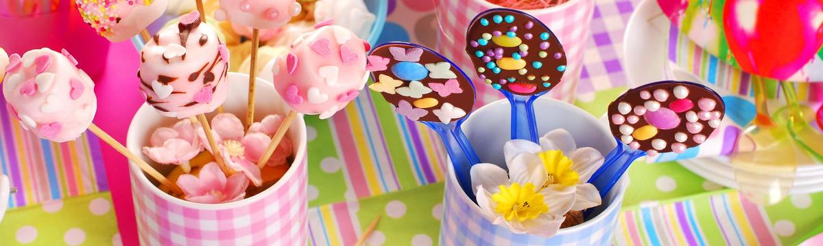 Colorful candy display with pink cake pops decorated with heart-shaped sprinkles and chocolate-covered spoons topped with bright sugar decorations, set on a festive, striped and polka-dotted tablecloth.