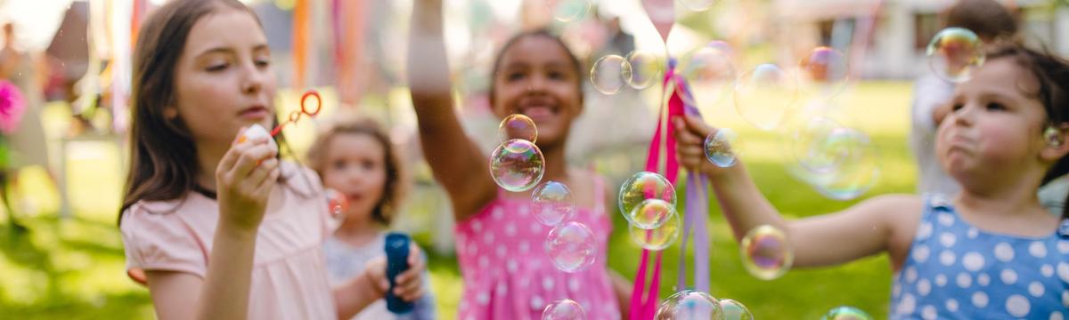 Children playing outdoors on a sunny day, blowing and chasing bubbles with big smiles and colorful clothing, surrounded by green grass and festive decorations.