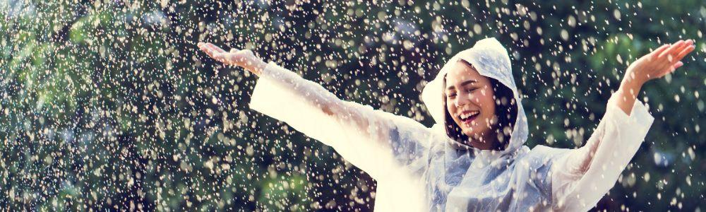 A young teenage girl wearing a clear rain-jacket is under the rain with a smile and open arms.  She is enjoying the moment and feeling of rain on her face because she is ready to have fun no matter the weather.  