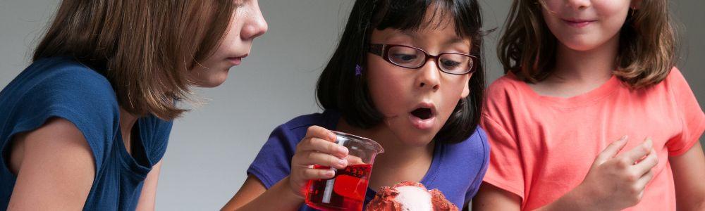 A group of 3 teenage girls making a volcano at home and ready to pour a red liquid from a beaker in order to trigger the lava reaction.