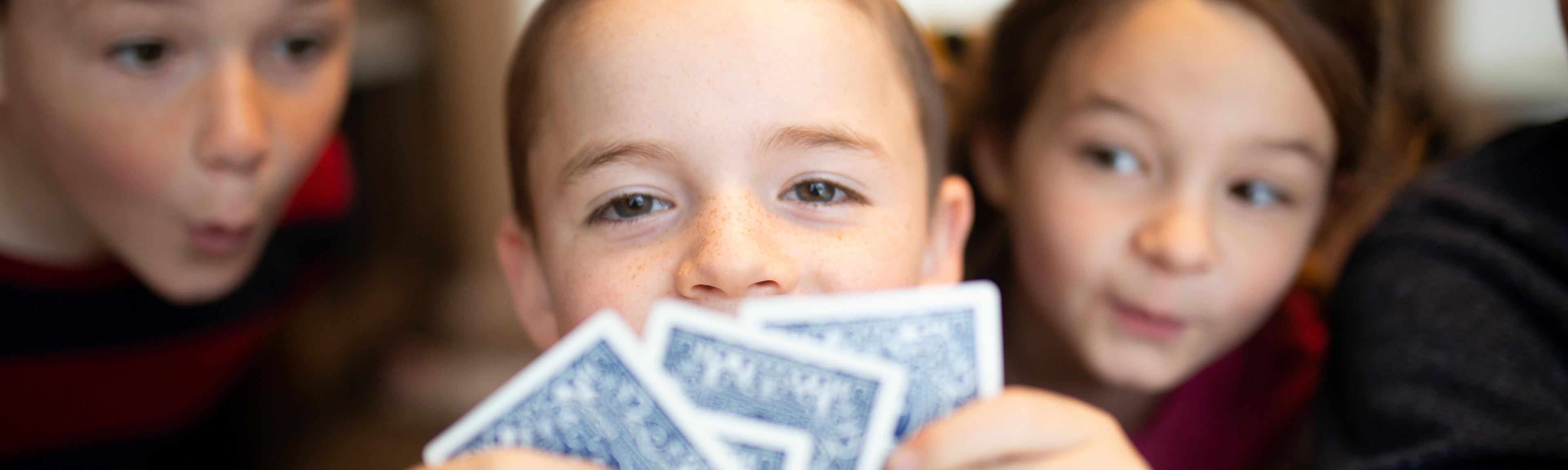 A pre-teen boy is making eye-contact with the camera while holding playing cards.  Two girls behind him are peaking in his cards.  It is a great pass-time activity during holidays that replaces screen time for kids, and encourages family time and social interaction.  