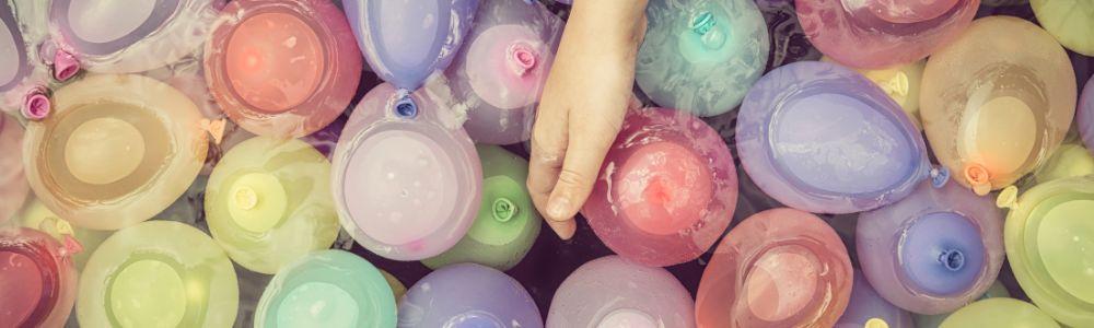 A child's hand dipping into a large bucket filled with water balloons and water, ready to play and perform sun experiments on a summer afternoon.  