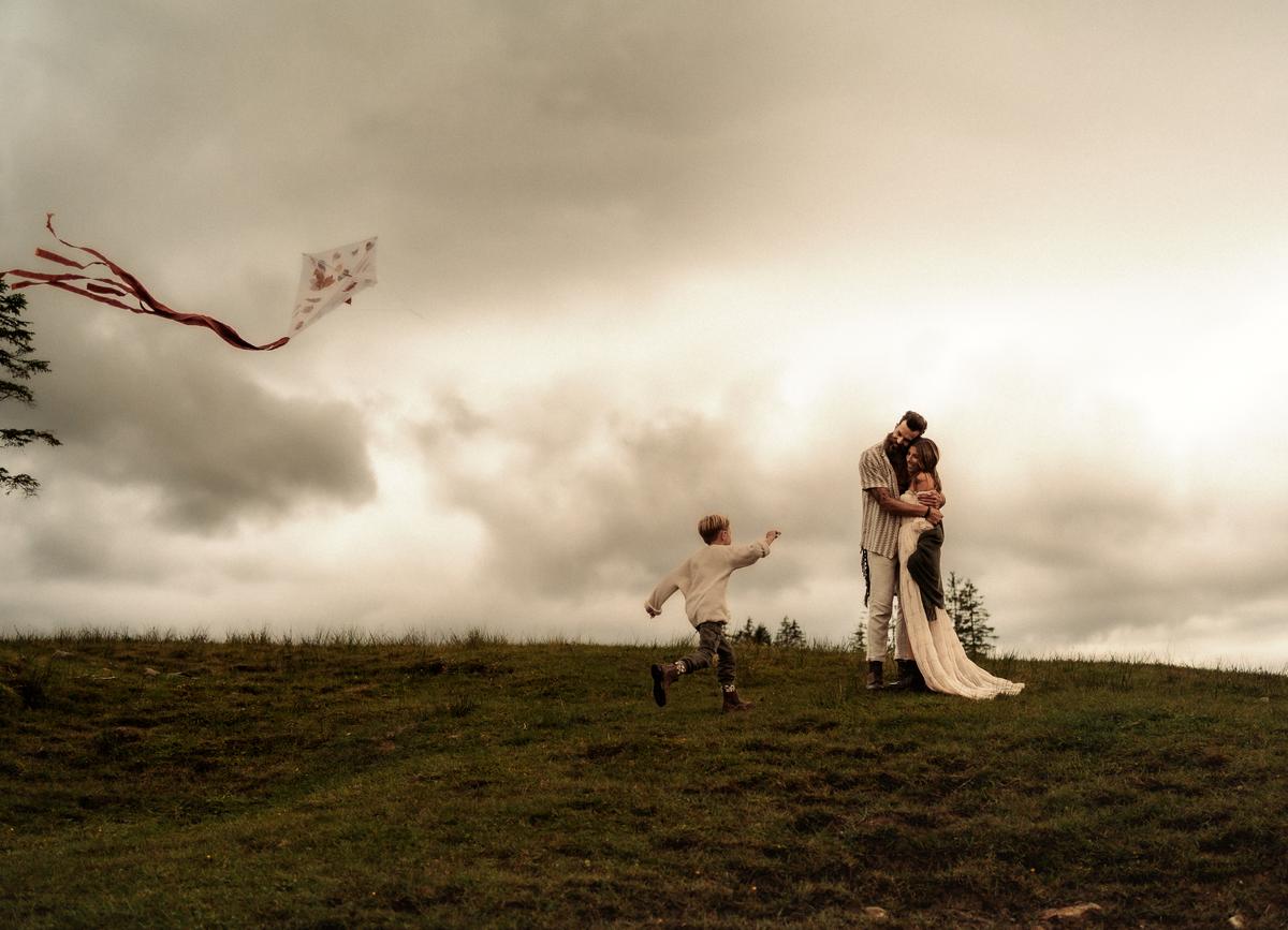 A family portrait in nature with a young boy running toward his parents who are embraced, in a windy nature, while a kite is flying on the left hand side.  Photographed by  family photographer Michèle Stucki.  