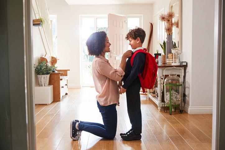 Mother and son in primary school age saying goodbye at the door.  She is fixing his collar and he is nervous while carrying a bright red backpack.