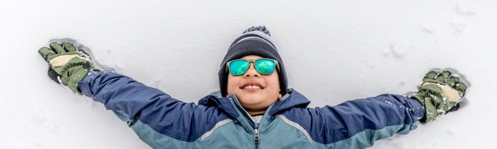 A young teenage boy with a big smile on his face looking up at the sky while lying in the snow with his hands wide open and enjoying himself after a long day doing winter sports.  He is wearing a wool hat and sunglasses.