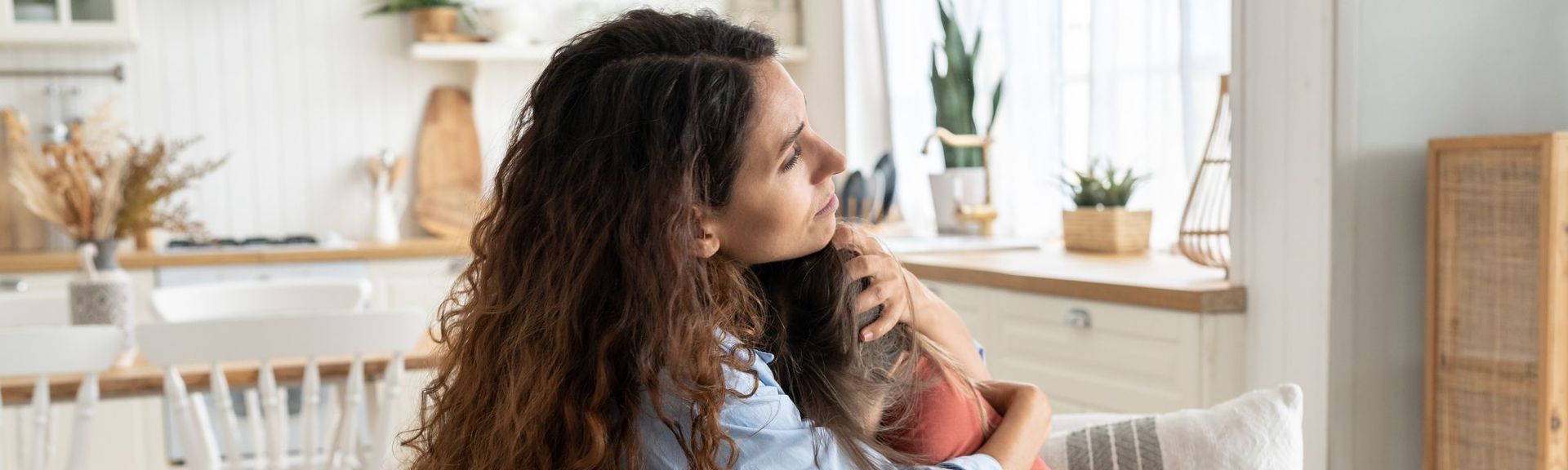 A white brown-haired mother embracing her pre-teen daughter on the sofa of their sunlit house, comforting her before her first day of school which is bringing her anxiety.  They are by the kitchen and can potentially have a comfort snack together.  