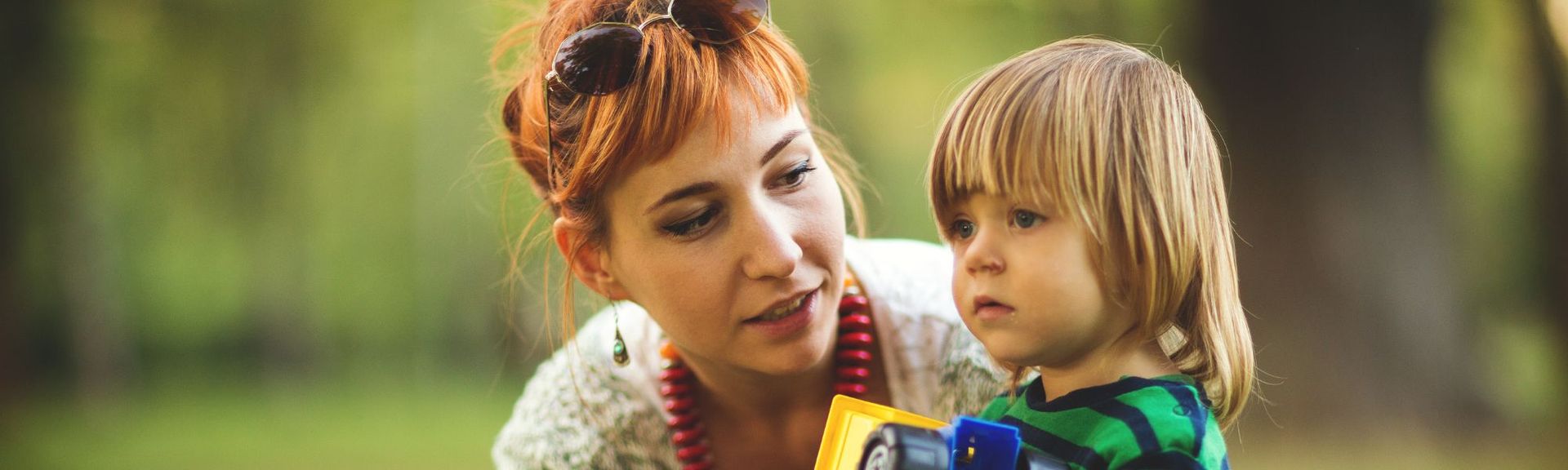 Mother spending quality time with her young child outdoors, holding toys and engaging in a caring conversation on a green, sunny day.