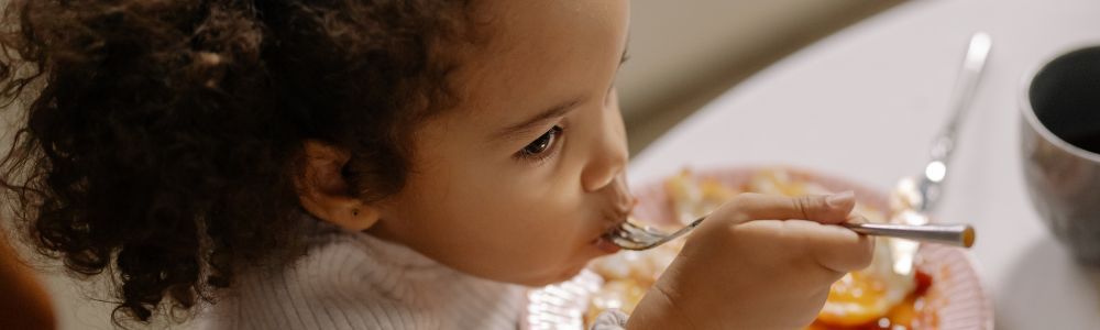 A toddler eating pancakes on a weekend at home, she is a colored girl with brown eyes enjoying her breakfast with her family nearby.