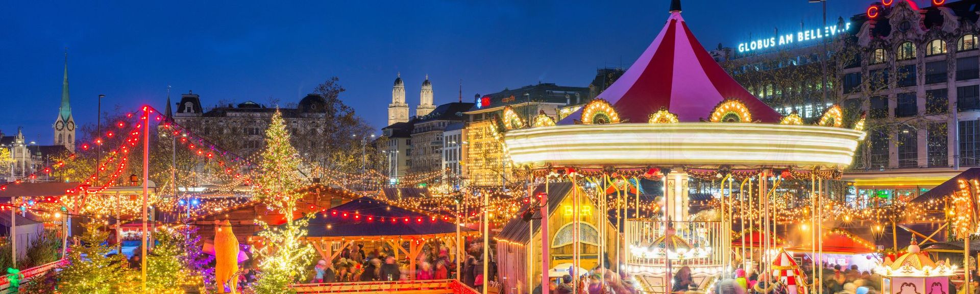 Abendstimmung am Zürcher Weihnachtsmarkt auf dem Sechseläutenplatz: Lichterketten, Karussell und Marktstände; Fraumünster- und Grossmünster-Türme im Hintergrund.