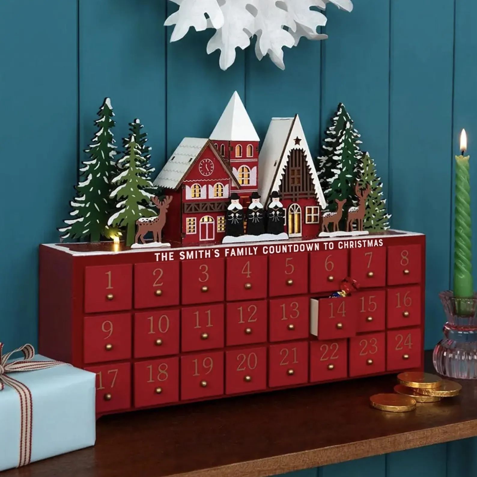 Personalisierbarer Holz-Adventskalender mit 24 roten Schublädchen und winterlichem Dorf obenauf – dekorativ und wiederverwendbar.