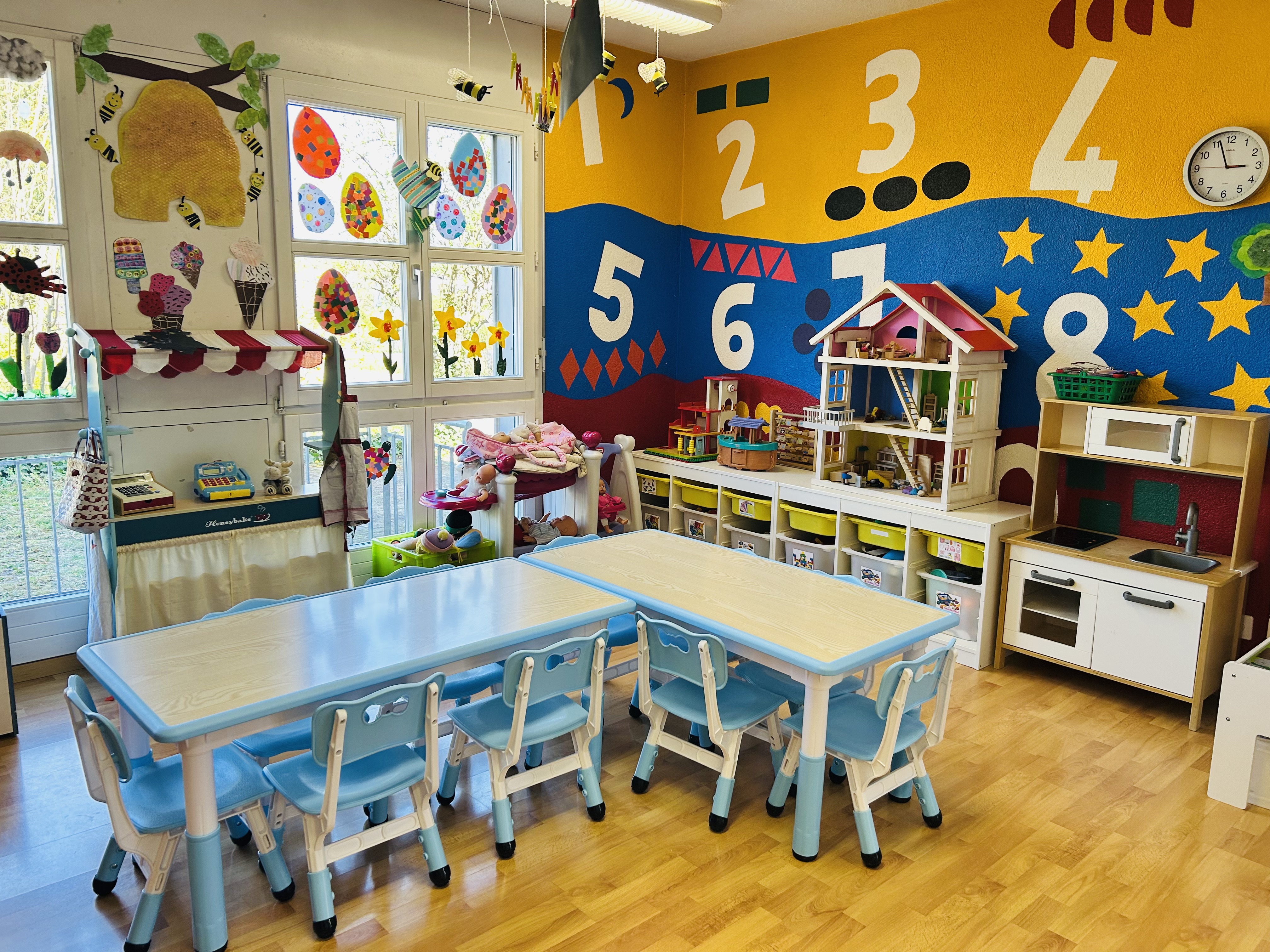 A colorful indoor area resembling a kindergarden, with yellow walls decorated with big posters of numbers and letters, balloons on the windows and white tables.  A child-friendly space where kids can learn through play and interactive games.  