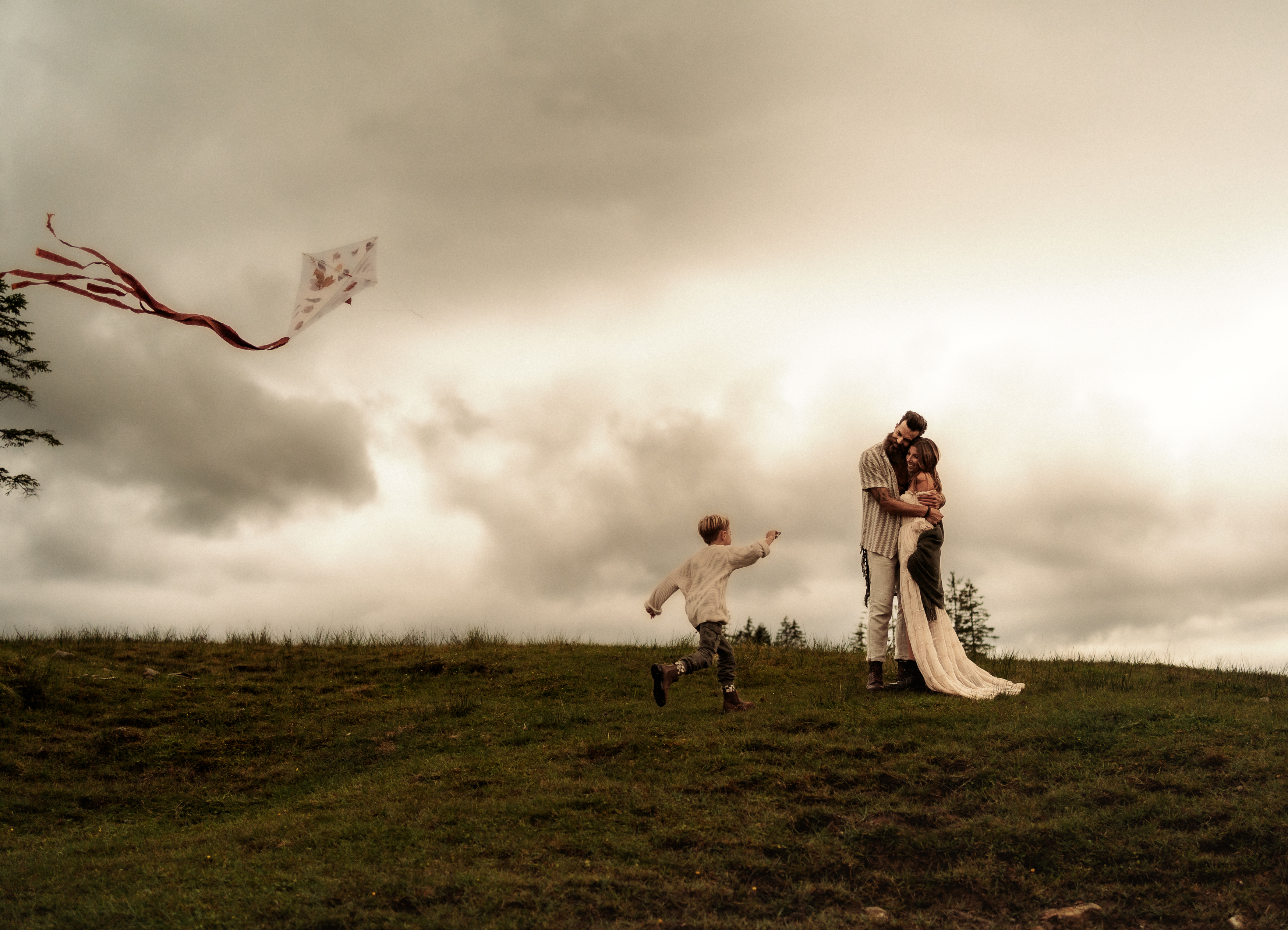 A family portrait in nature with a young boy running toward his parents who are embraced, in a windy nature, while a kite is flying on the left hand side.  Photographed by  family photographer Michèle Stucki.  